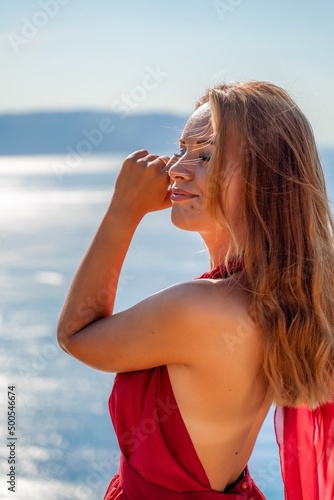 Smiling young woman in a red dress looks at the camera. A beautiful tanned girl enjoys her summer holidays at the sea. Portrait of a stylish carefree woman laughing at the ocean.