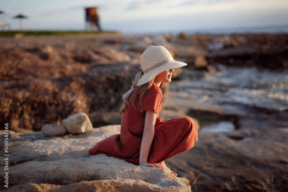 Cute European teenage girl in hat and dress on the rocks on an empty seashore,