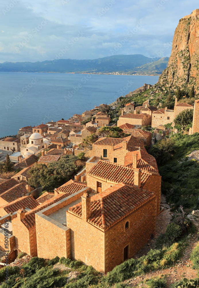 View of fortified medieval castle Monemvasia with Aegean sea and coast as background. Stone houses with dusty pink terracotta tiled roofs and blue sea.