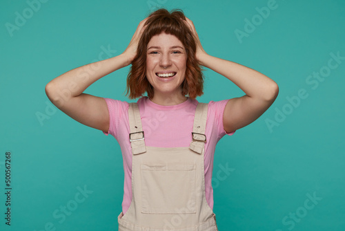 indoor portrait of young ginger female posing over blue background touching her hair with broad smile on her face