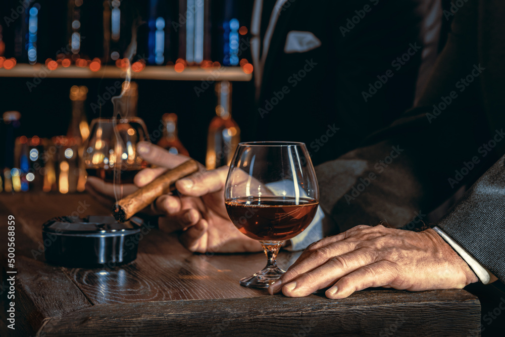 Man's hands with a cigar, elegant glass of brandy on the bar counter. Alcoholic drinks, cognac, whiskey, port, brandy, rum, scotch, bourbon. Vintage wooden table in a pub at night