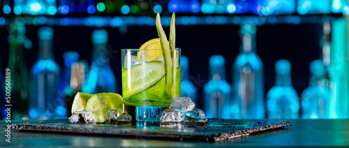 Cocktail with cucumber, ice on bar counter in a restaurant, pub. Fresh tonic drink with lime juice, mint, gin, cucumber juice. Alcoholic cooler beverage at nightclub on dark background photo