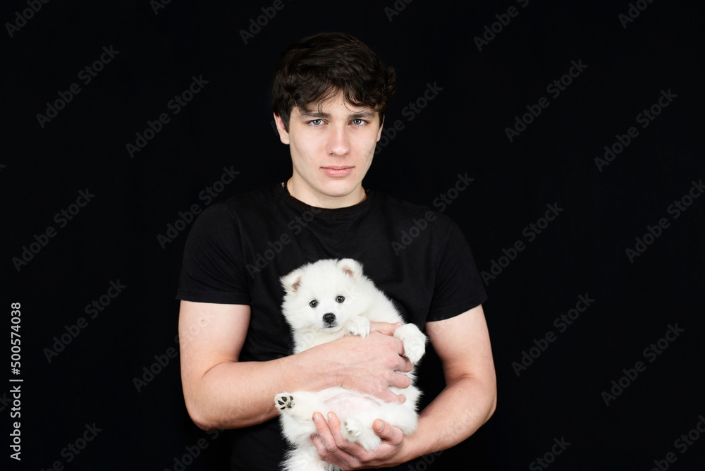 Young teen boy on a dark background in a dark T-shirt with a white dog puppy in his arms. The boy still does not know how to raise a puppy and is a little confused Young teen boy on a dark background in a dark T-shirt with a white dog puppy in his arms. The boy still does not know how to raise a puppy and is a little confused