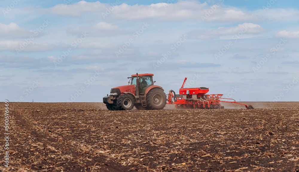 Fototapeta premium Spring sowing season. Farmer with a tractor sows corn seeds on his field. Planting corn with trailed planter. Farming seeding. The concept of agriculture and agricultural machinery.