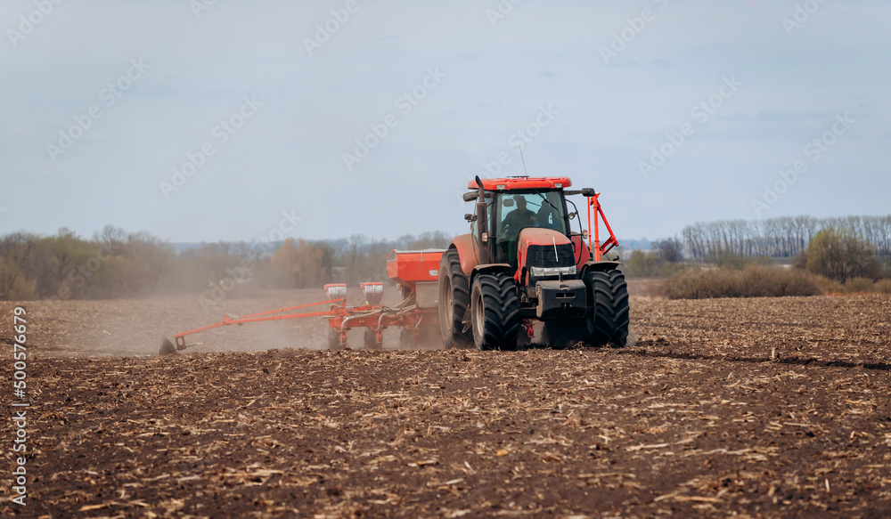 Fototapeta premium Spring sowing season. Farmer with a tractor sows corn seeds on his field. Planting corn with trailed planter. Farming seeding. The concept of agriculture and agricultural machinery.