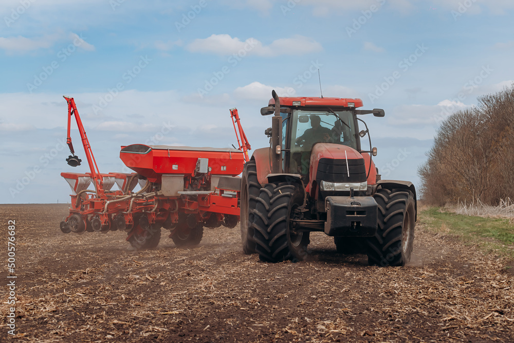 Fototapeta premium Spring sowing season. Farmer with a tractor sows corn seeds on his field. Planting corn with trailed planter. Farming seeding. The concept of agriculture and agricultural machinery.