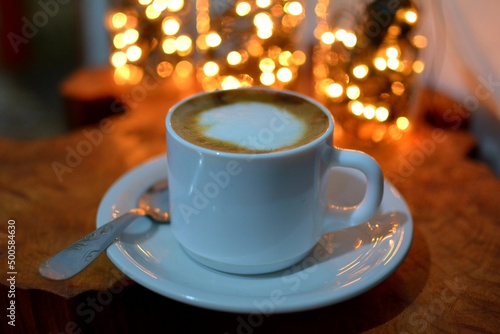 taza de café blanca, disfrutando de un buen late en un día frío sobre una mesa rústica con luces de fondo.