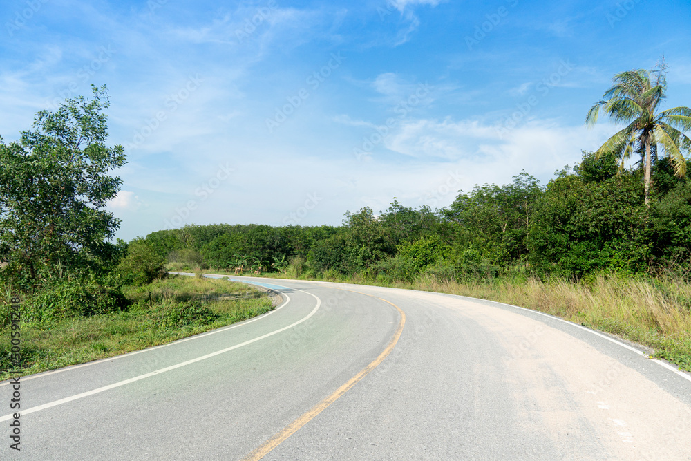 Naklejka premium curving path leading forward of the asphalt road has sand on top of the road in Thailand. beside ground of green grass and trees. Under the blue sky.