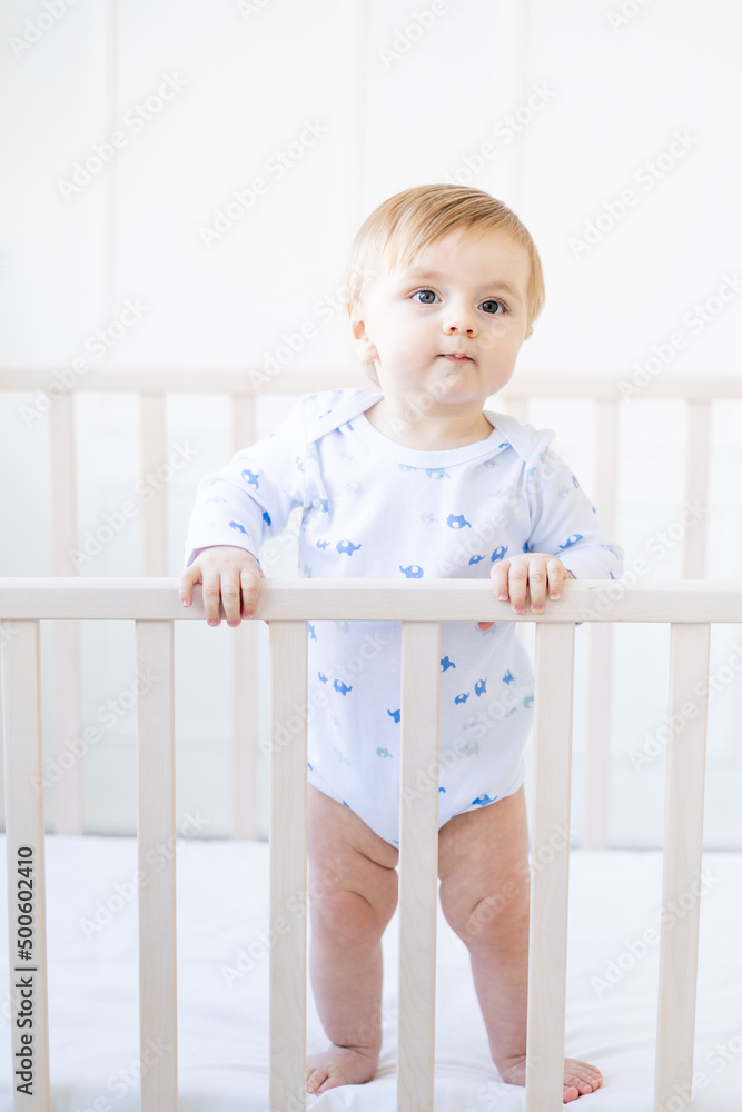 a blonde baby boy in a crib at home with blue and white cotton bedding stands holding on to the side and smiles in the morning, the concept of children's goods and accessories a blonde baby boy in a crib at home with blue and white cotton bedding stands holding on to the side and smiles in the morning, the concept of children's goods and accessories