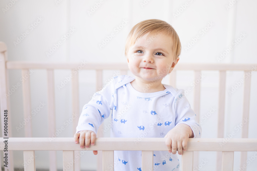 smiling blonde baby boy in a crib at home with blue and white cotton bedding stands holding on to the side and laughs in the morning, the concept of children's goods and accessories smiling blonde baby boy in a crib at home with blue and white cotton bedding stands holding on to the side and laughs in the morning, the concept of children's goods and accessories