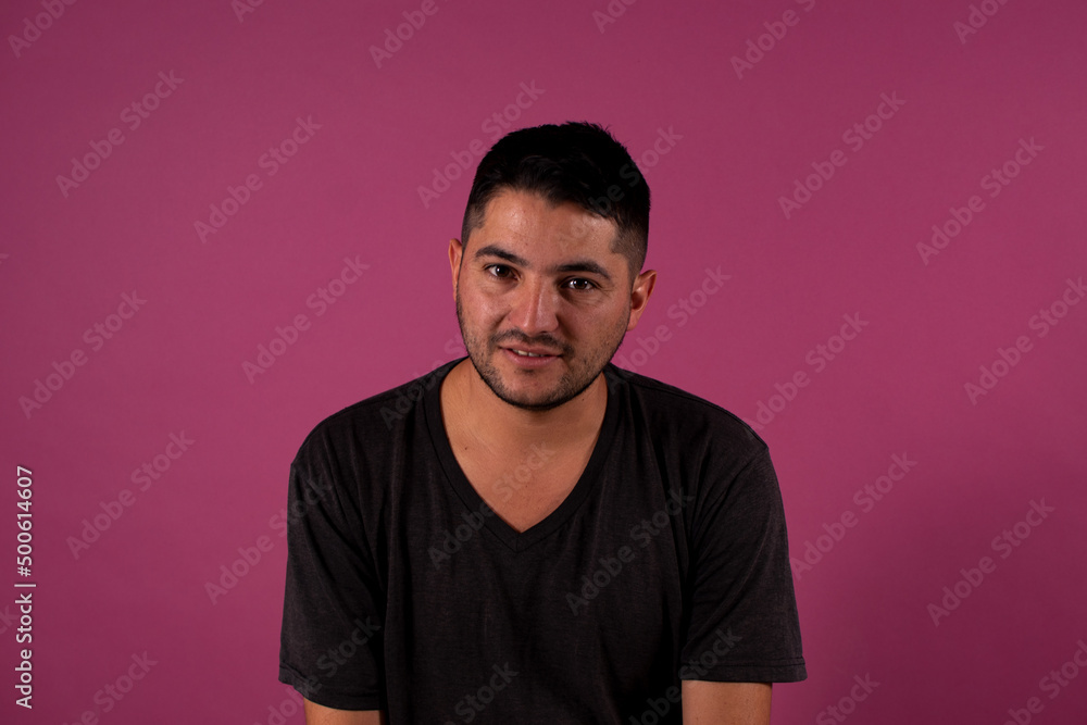 Obraz premium looking at the camera with a shy smile , young brunet man, 20s, wears a blue shirt looking at camera smiling isolated on light pink background studio portrait. People emotions lifestyle concept