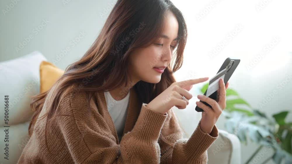 Happy young asian woman holding credit card using instant mobile payments at home. Smiling Female customer shopper making purchase on smartphone receiving cash back concept. E-banking app service.	