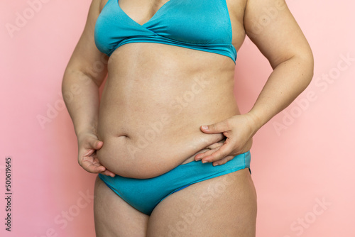 Pinch woman sagging belly with fingers closeup, folds on stomach, loose skin and cellulite. Naked overweight plus size girl on pink background in blue underwear. Concept of dieting and body control.