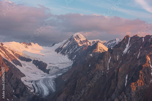 Scenic aerial view to high snow mountains in early morning at dawn. Awesome scenery with sunlit gold pinnacle in cloudy sky at sunrise. Morning landscape with snow mountain peak in golden sunlight.