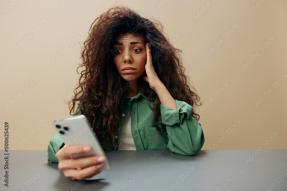 Confused sad unhappy tanned curly Latin lady read fake news sit at the table with phone isolated over pastel beige background look at screen. Copy space Mockup Banner. Shock Information from call