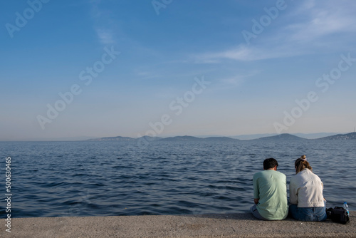 Back view of young couple sitting by the sea on beach. Desire, hope to go far away. Making holiday plan. Minimal Concept. Free area for text or ads © enver