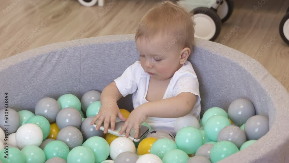 Kid playing with smartphone in ball pit at home. Happy kid lying in soft ball pit pool with colorful balls for toddlers, caucasian baby boy having fun playing at home. . High quality 4k footage Kid playing with smartphone in ball pit at home. Happy kid lying in soft ball pit pool with colorful balls for toddlers, caucasian baby boy having fun playing at home. . High quality 4k footage
