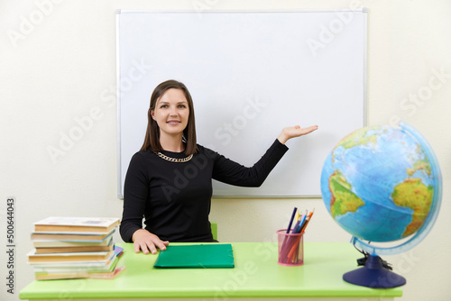 Young teacher sitting at a table near empty white board in classroom photo