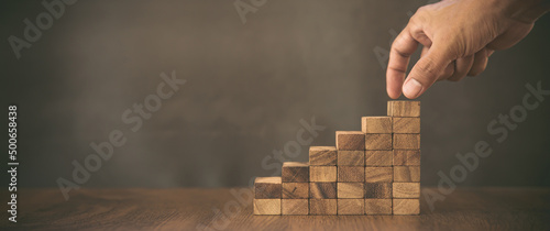 Fotografie, Obraz Hand is placing wood block tower stack in pyramid stair step with caution to prevent collapse or crash concepts of financial risk management and strategic planning and business challenge plan