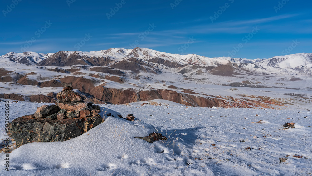 A picturesque mountain range against the blue sky. The red-brown slopes are covered with snow. In the foreground is a pyramid of stones. Dry grass in snowdrifts. Altai. Kyzyl-Chin