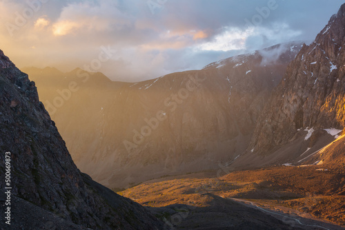 Scenic alpine landscape with mountain wall in low clouds in golden sunrise colors. Colorful view to high mountains and rocks in gold morning sunlight. Golden light in mountain valley at early morning.