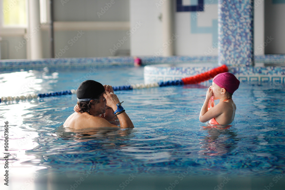Happy kid toddler learns to swim with coach in pool, daddy grandpa teaches kids to swim and dive in pink swimming cap