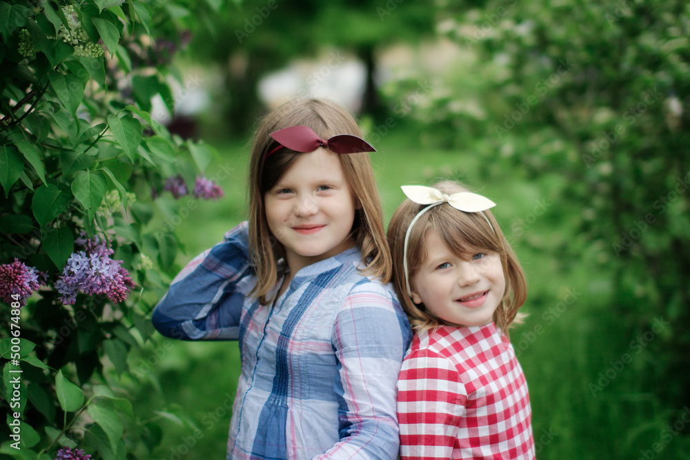 Cute sibling sister girls with a headband with a bow are hugging among green Cute sibling sister girls with a headband with a bow are hugging among green
