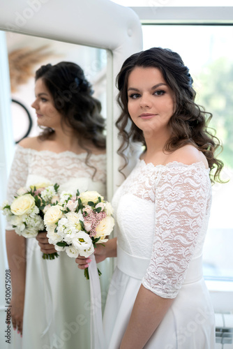 A beautiful bride with curly black hair in a white wedding dress with lace sleeves and a bouquet of white flowers in her hands in the wedding salon is reflected in the mirror