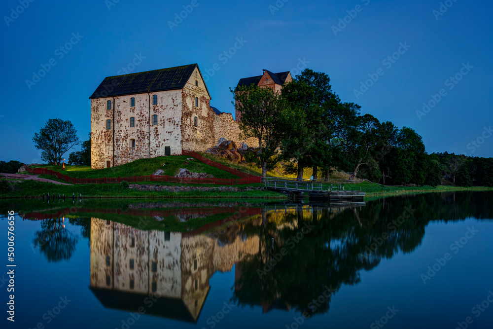 Lit medieval Kastelholm Castle and its reflections on a calm river in Åland Islands, Finland, at dusk in the summer.