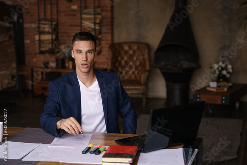Young man in jacket working from home doing paperwork using laptop and holding a pen in his hand while sitting at his desk. Performs work remotely
