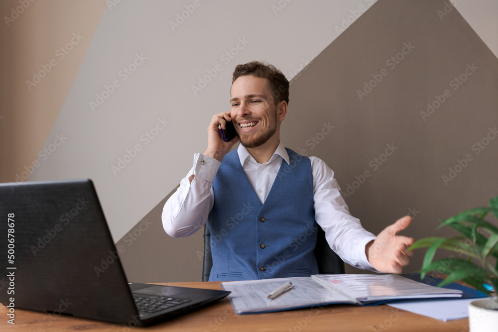 Portrait smiling business man with beard talking on his smartphone while working on his computer while talking, sitting in his office wearing shirt and jacket. Negotiates or works with client at work Portrait smiling business man with beard talking on his smartphone while working on his computer while talking, sitting in his office wearing shirt and jacket. Negotiates or works with client at work
