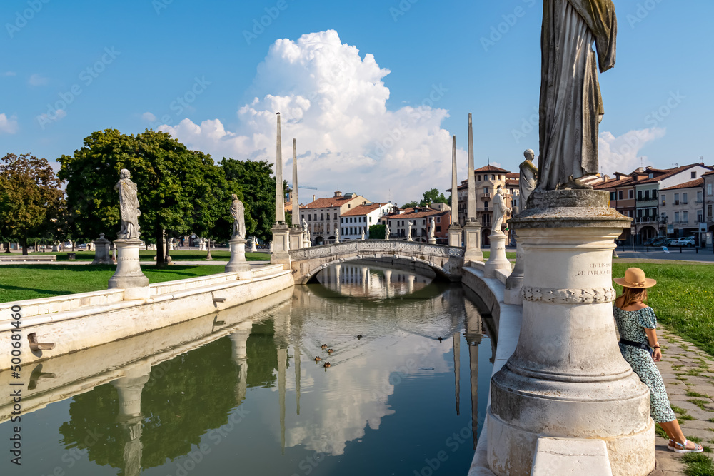Naklejka premium Woman in a dress and hat leaning against columns of a statue at Prato della Valle, square in the city of Padua, Veneto, Italy, Europe. Isola Memmia surrounded by canal bordered by two rings of statues