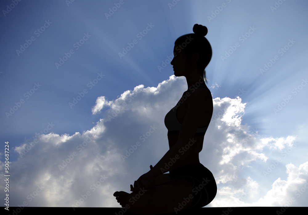 Woman side silhouette in Yoga Easy Meditating pose with rays of sunlight through Cloudy sky
