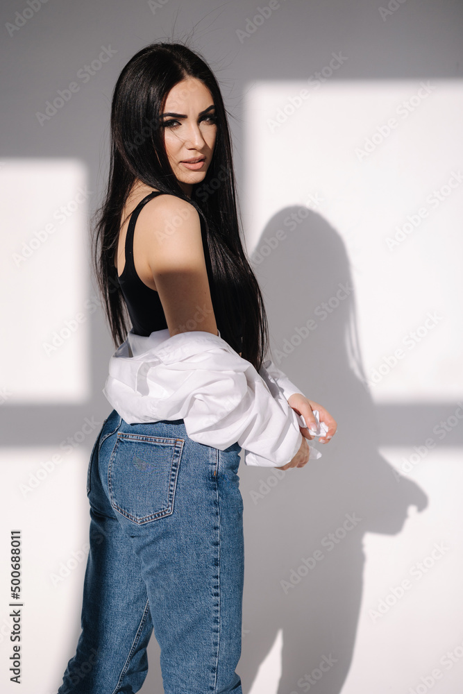 Fotografia do Stock: Pretty brunette hair woman in blue jeans and white  shirt standing in front of white wall. Shadow on white background | Adobe  Stock