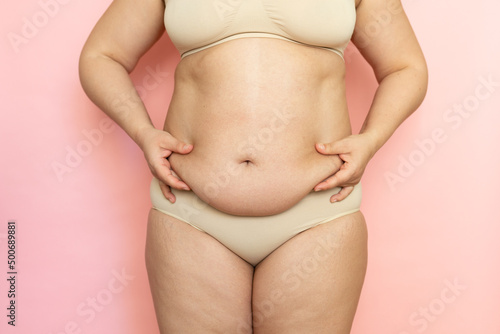Measure woman sagging belly closeup, folds on stomach, loose skin and cellulite, obesity. Naked overweight plus size girl on pink background in beige underwear. Concept of dieting and body control.