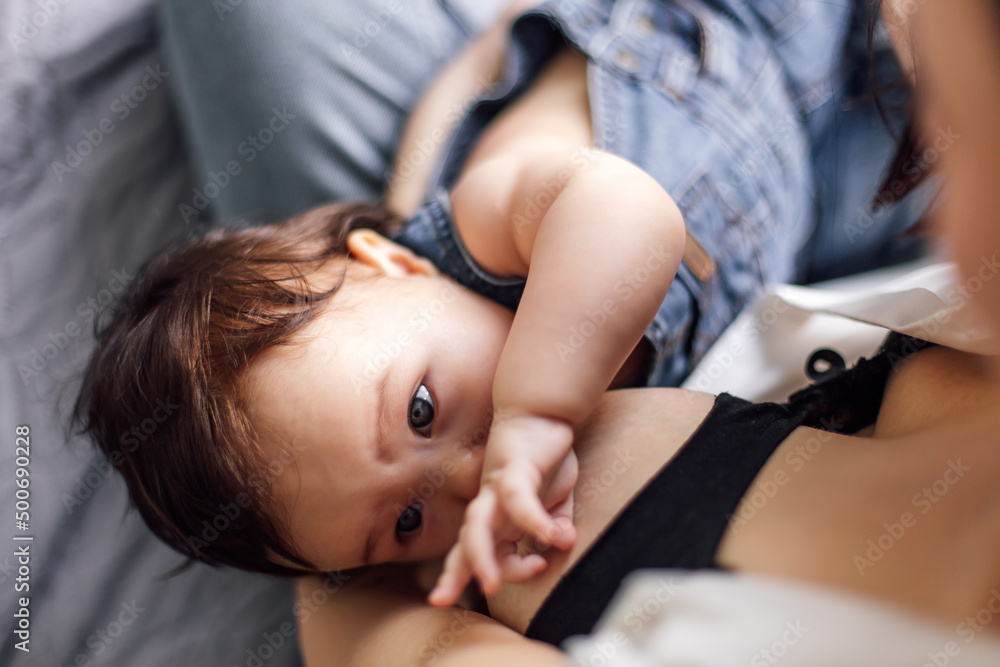 Closeup portrait of breastfeeding little girl in denim baby romper, gray blurred background. Baby drink breast milk and get nutrients for healthy growth. Concept of maternal affection and childcare. Closeup portrait of breastfeeding little girl in denim baby romper, gray blurred background. Baby drink breast milk and get nutrients for healthy growth. Concept of maternal affection and childcare.