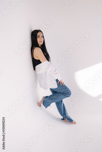Pretty woman barefoot in studio. Beautiful brunette girl in black topic with white shirt and blue denim jeans. White background of cyclorama
