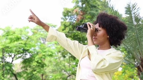 Young Africa American woman looking natural and using binoculars in public park with a happy face standing and smiling. Concept of nature and learning.