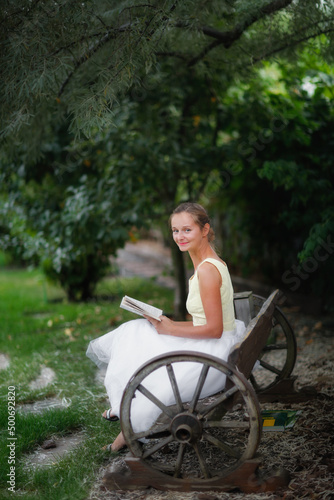 Cute european girl woman with a book on a garden bench under a willow tree, a beautiful garden and a wooden bench made of wagon wheels, landscape art and a beautiful well-groomed garden