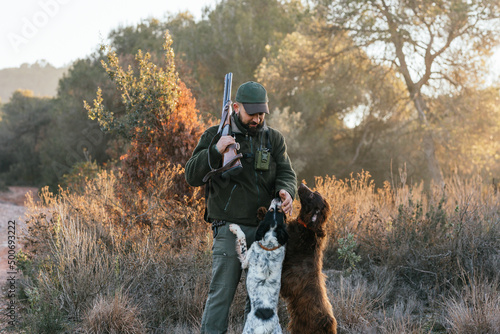 Hunter with his shotgun on shoulder petting his two dogs in nature