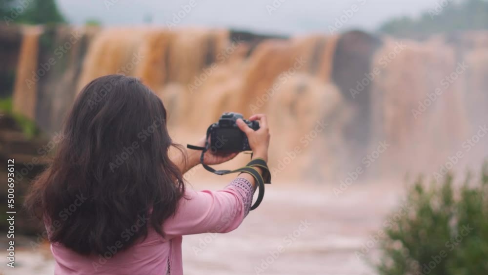 Indian young woman taking photographs with her dslr in front of the Gira Waterfall while travelling at Saputara in Gujarat, India. Female travel vlogger shooting with camera Indian young woman taking photographs with her dslr in front of the Gira Waterfall while travelling at Saputara in Gujarat, India. Female travel vlogger shooting with camera