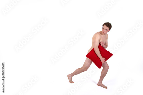 A handsome young man without clothes bashfully covers himself with red pillows. Topless guy isolate.