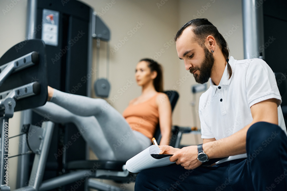 Obraz premium Fitness instructor taking notes while athletic woman is exercising in the background.
