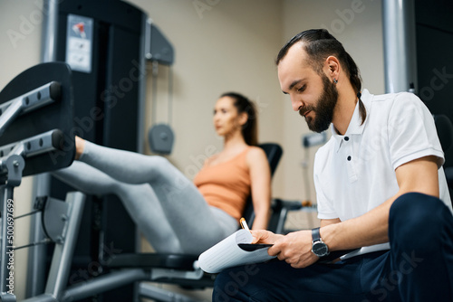 Fitness instructor taking notes while athletic woman is exercising in the background.