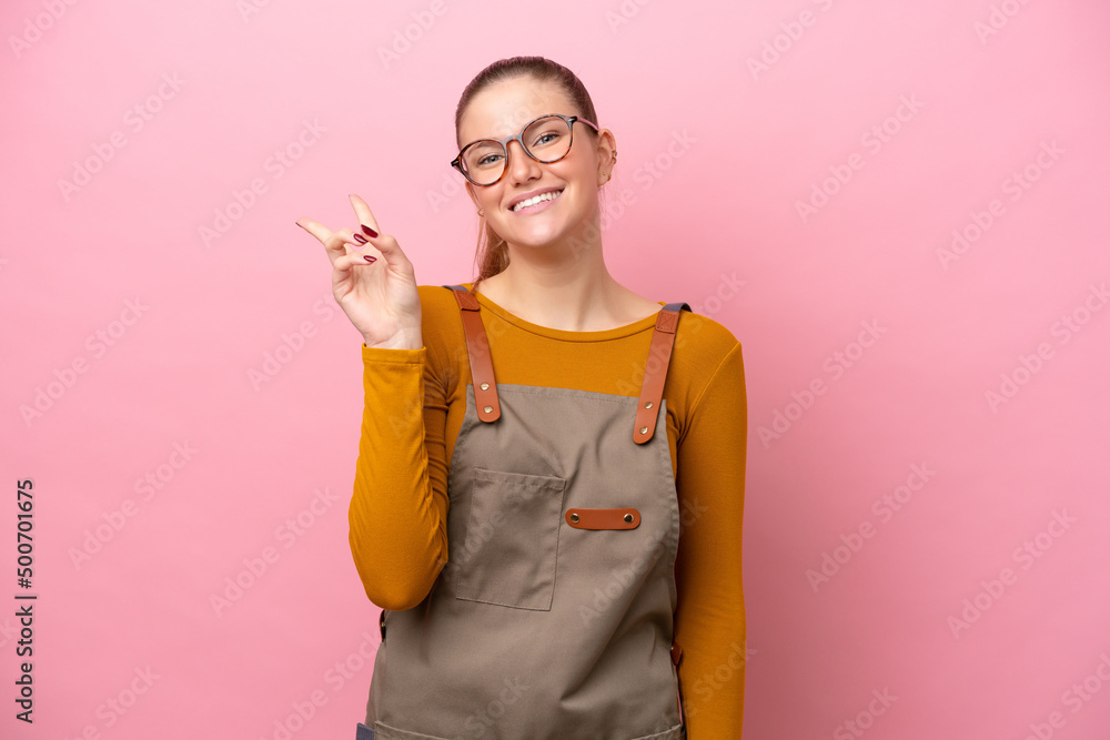 Woman with apron isolated on pink background smiling and showing victory sign Woman with apron isolated on pink background smiling and showing victory sign