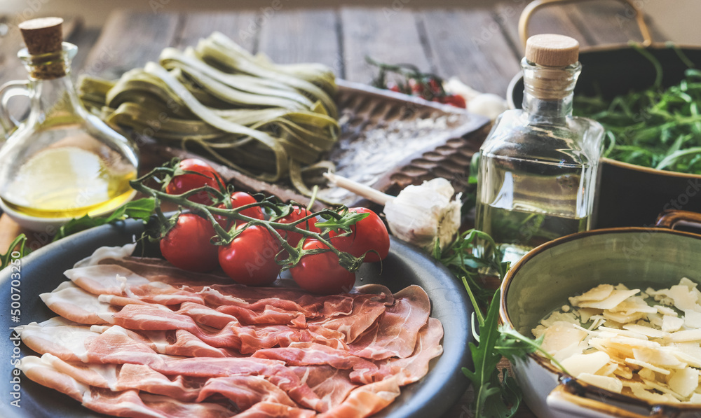Italian food background with ham, tomatoes, green pasta, Parmesan cheese, olive oil and arugula at rustic wooden kitchen table. Cooking preparation with traditional Mediterranean food. Top view