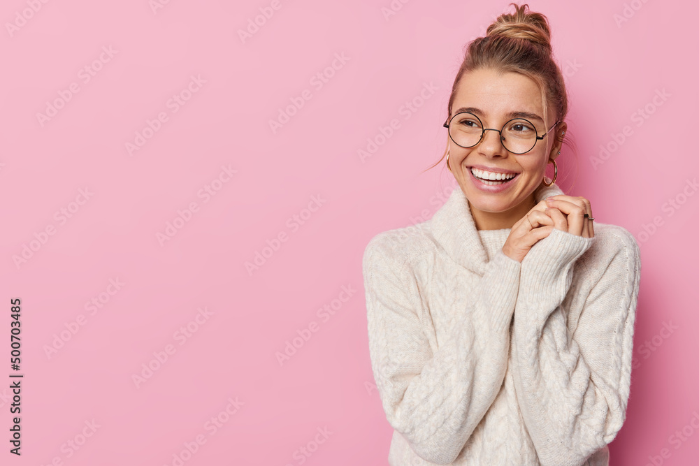 Studio shot of lovely cheerful woman keeps hands together smiles broadly looks away feels satisfied wears knitted sweater isolated over pink background empty space for your promotional content Studio shot of lovely cheerful woman keeps hands together smiles broadly looks away feels satisfied wears knitted sweater isolated over pink background empty space for your promotional content