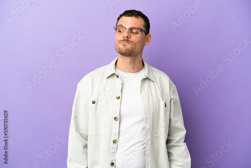 Brazilian man over isolated purple background having doubts while looking side © luismolinero