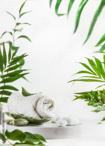 Spa setting with towel, jade massage roller and stones on white podium at white background with tropical leaves. Modern skin care treatment . Beauty products. Front view.