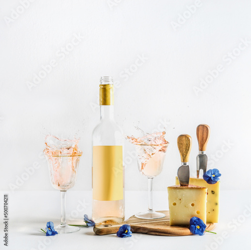 Wine and cheese setting with splashing in wineglasses and wine bottle with empty label mock up, cheese knifes, cutting board and blue flowers. Front view.